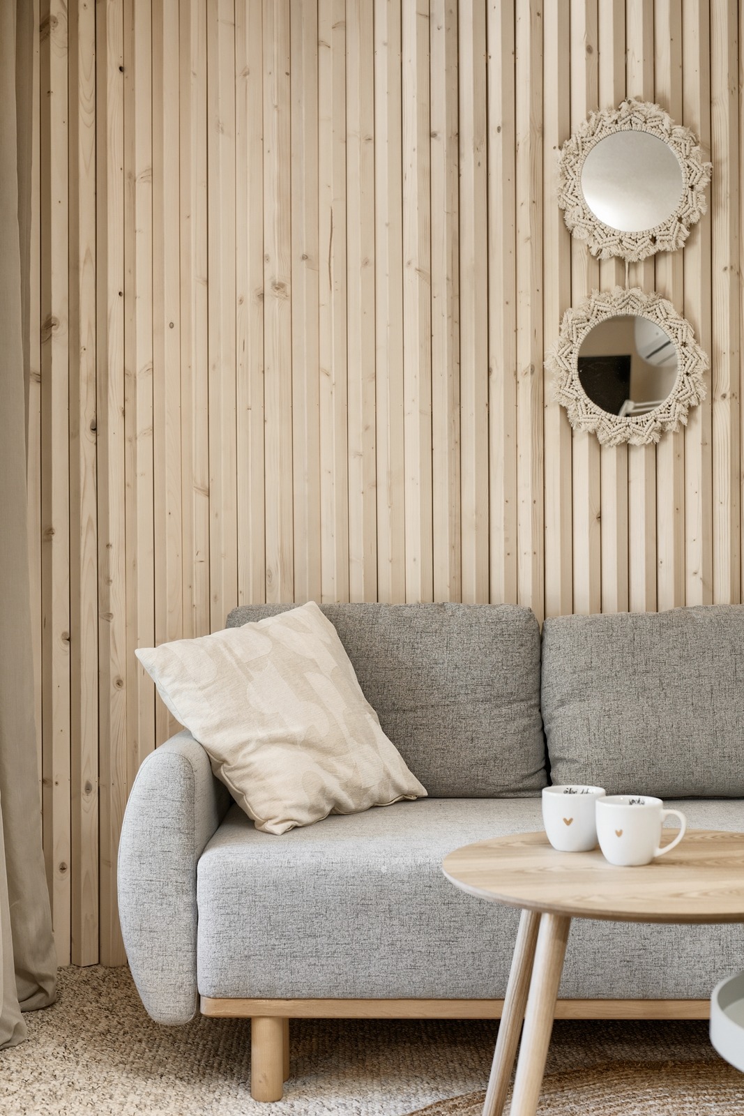 Szary narożnik z poduszkami i okrągły stolik z dwoma kubkami z serduszkami na tle ściany z drewnianych paneli i dwóch okrągłych luster z makramą.