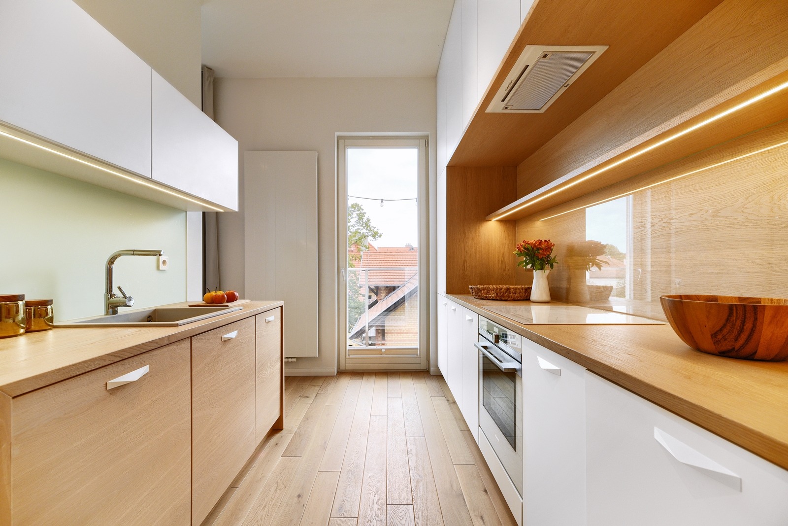 Jasna kuchnia z drewnianymi blatami i białymi szafkami, oświetlenie LED pod szafkami, drewniana podłoga, okno z widokiem na domy.