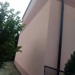 Różowa elewacja budynku z widoczną rynną spustową i fragmentem ogrodzenia, zieleń drzew w tle.