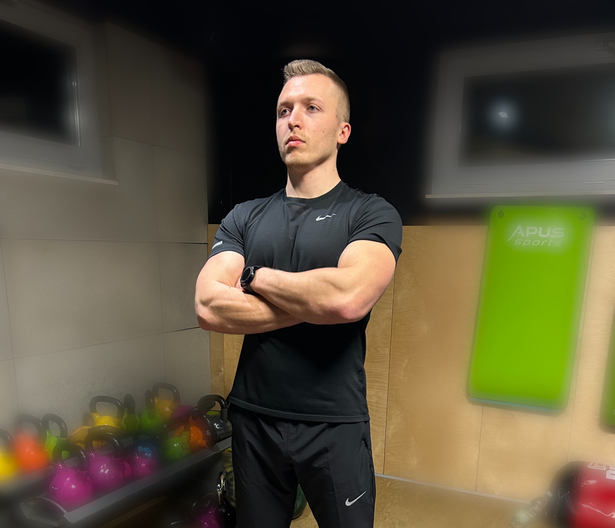 Młody mężczyzna w sportowym stroju, z założonymi rękami, stojący na tle siłowni z kettlebells i matami do ćwiczeń.