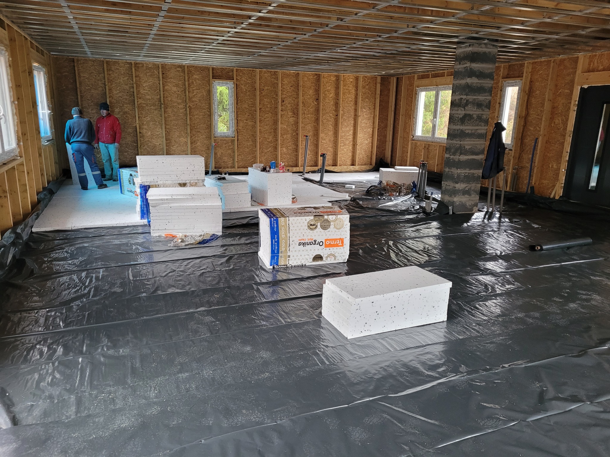 Wnętrze w trakcie prac przygotowawczych pod ocieplenie podłogi: rozłożona czarna folia, styropianowe płyty termoizolacyjne w opakowaniach i luzem, widoczni dwaj pracownicy w oddali, surowa...