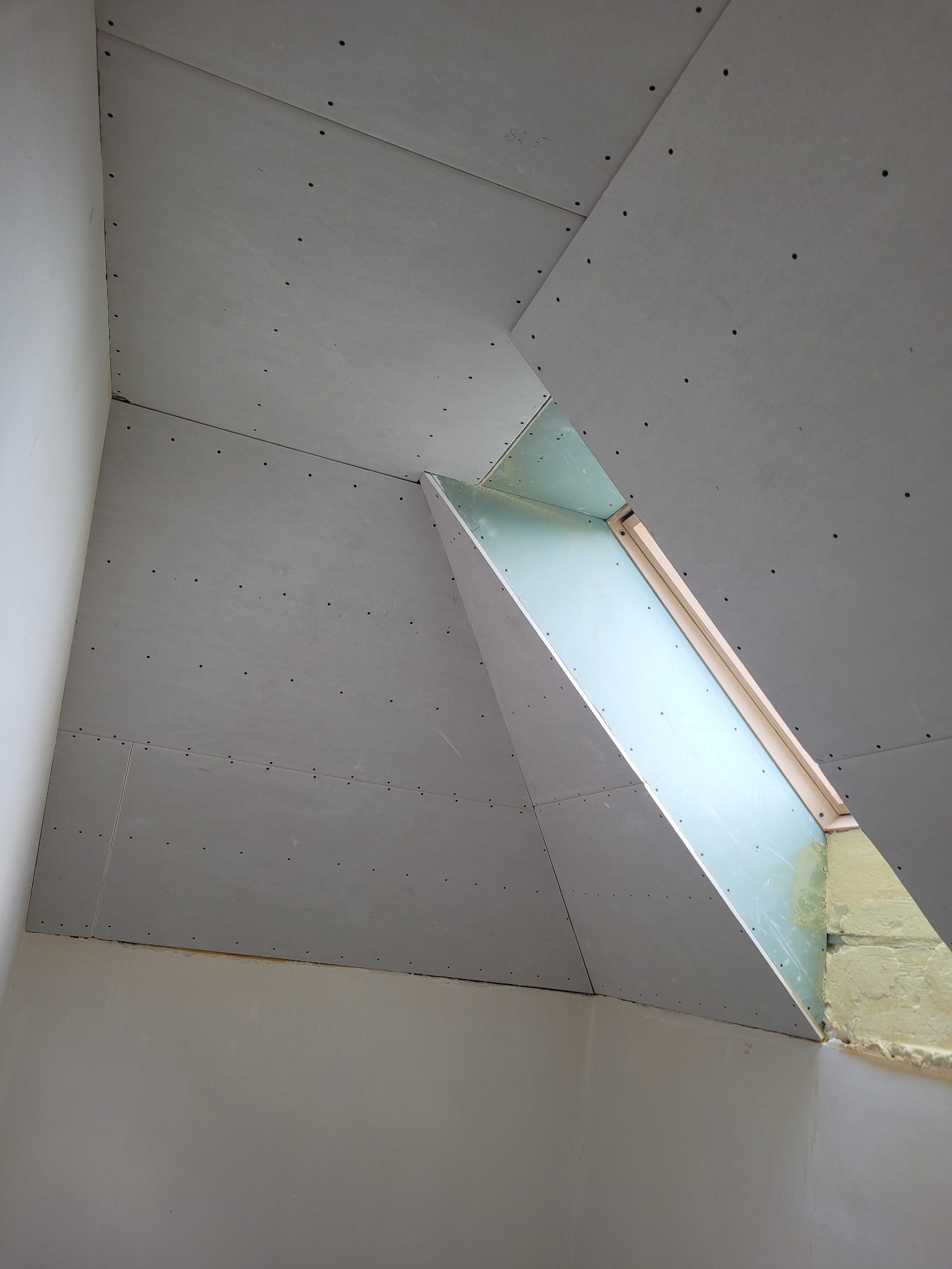 Skośny sufit poddasza z zamontowaną płytą karton-gips wokół okna dachowego, widoczne wkręty montażowe i szczelina okienna z izolacją.