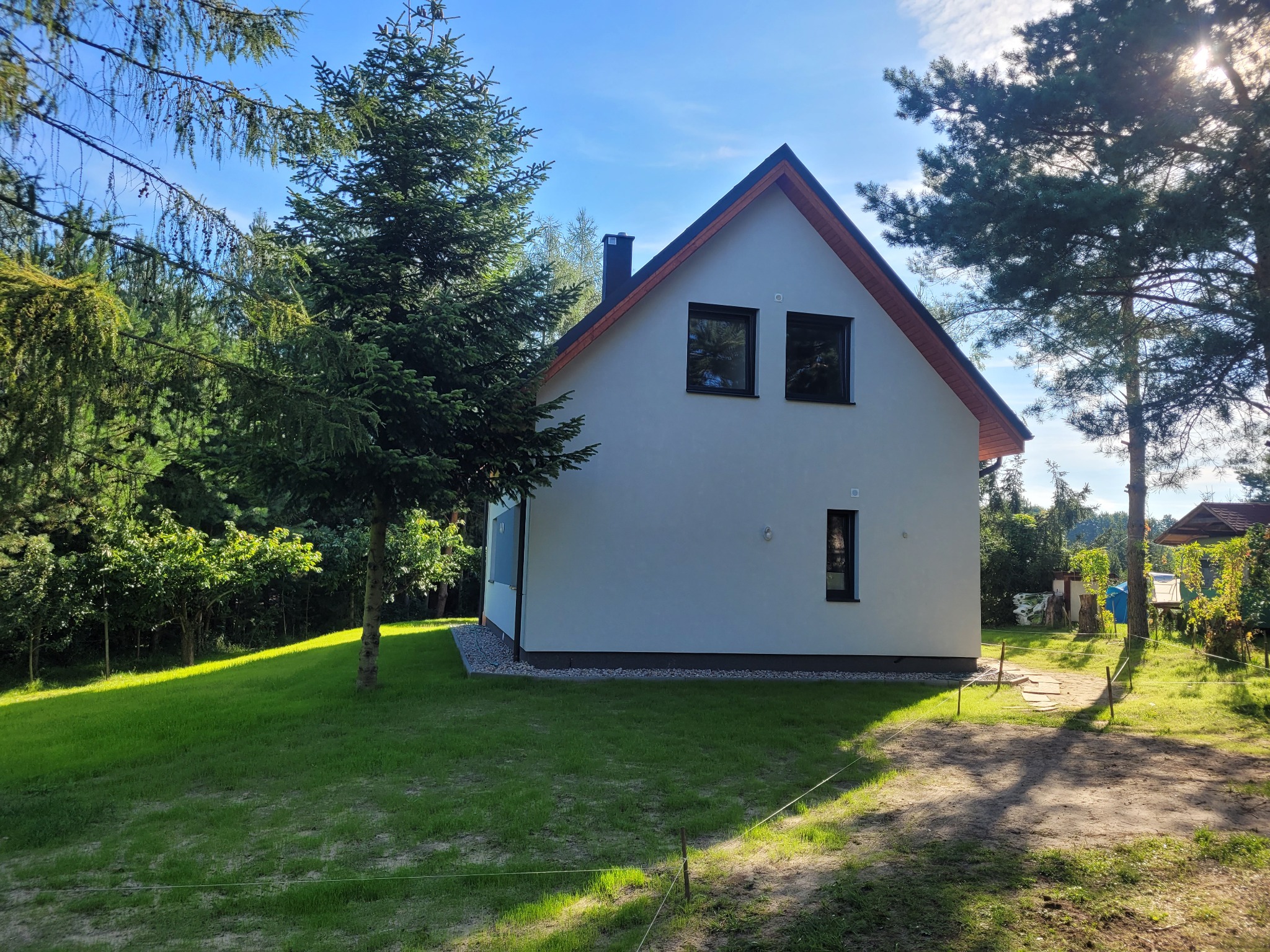 Świeżo otynkowany, biały dom jednorodzinny z ciemną dachówką i oknami, otoczony zielonym trawnikiem i drzewami w słoneczny dzień.