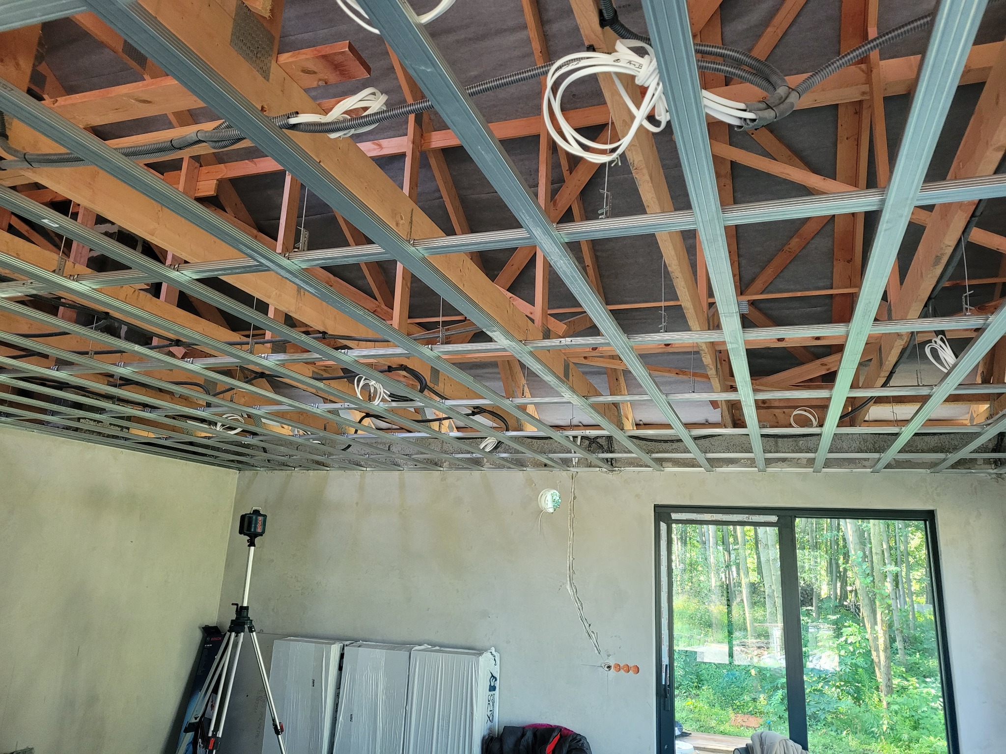 Konstrukcja sufitu podwieszanego z płyt kartonowo-gipsowych, widoczne drewniane belki stropowe, metalowy stelaż, przewody elektryczne oraz laser krzyżowy na statywie w pomieszczeniu z dużym oknem.