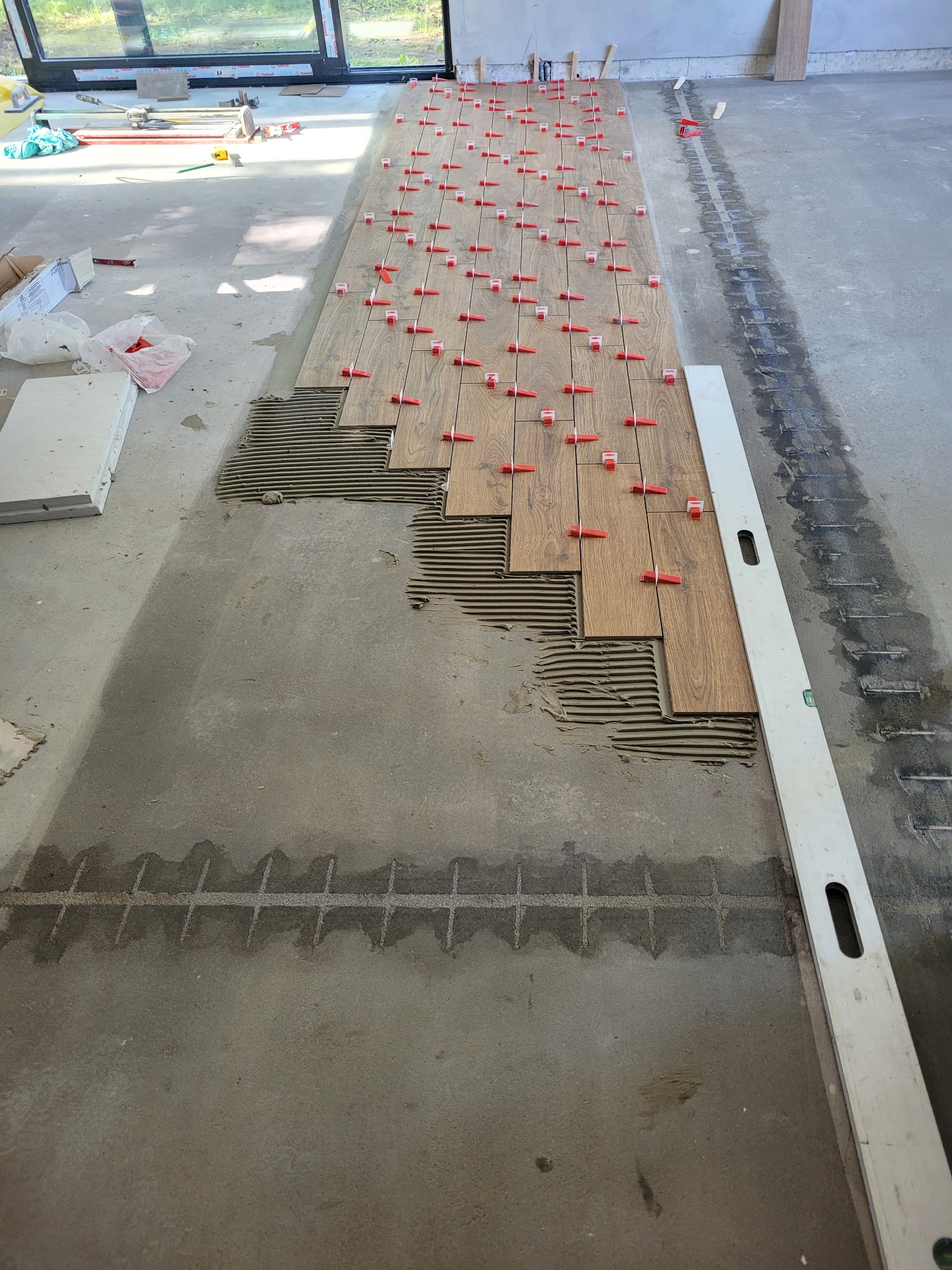 Układanie podłogi z drewnopodobnych płytek, widoczne rzędy płytek z czerwonymi klipsami poziomującymi, resztki kleju i poziomica na podłodze.