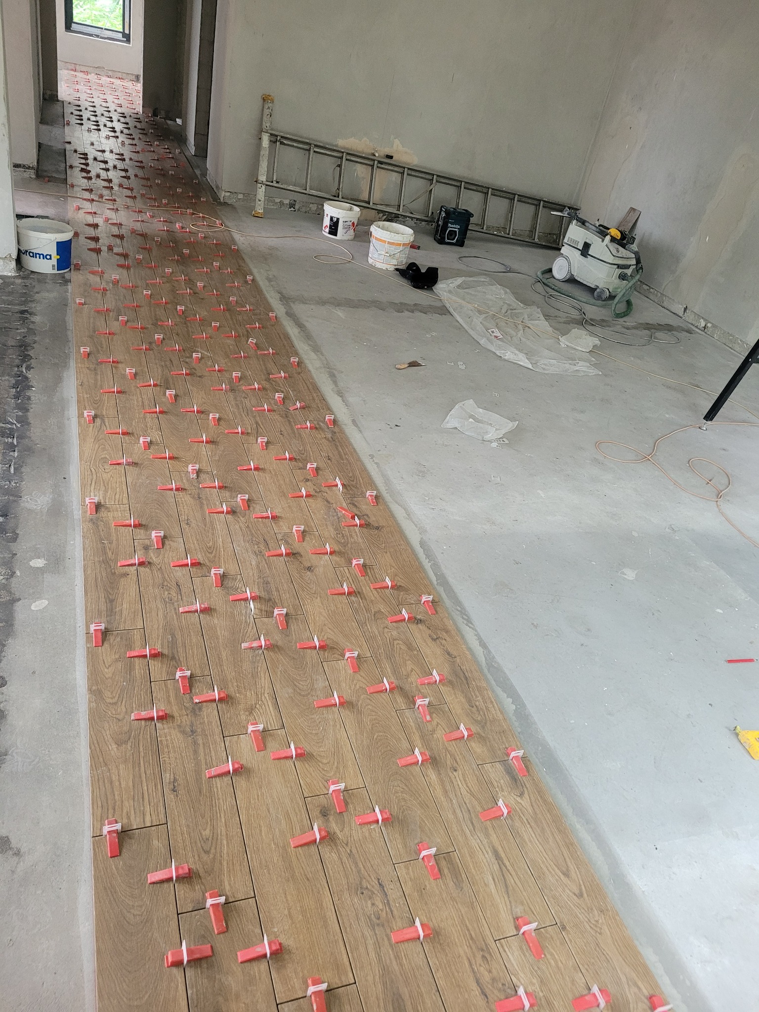 Proces układania paneli podłogowych imitujących drewno, z użyciem czerwonych klinów dystansowych, widok na długi korytarz w surowym stanie, z drabiną i odkurzaczem przemysłowym w tle.