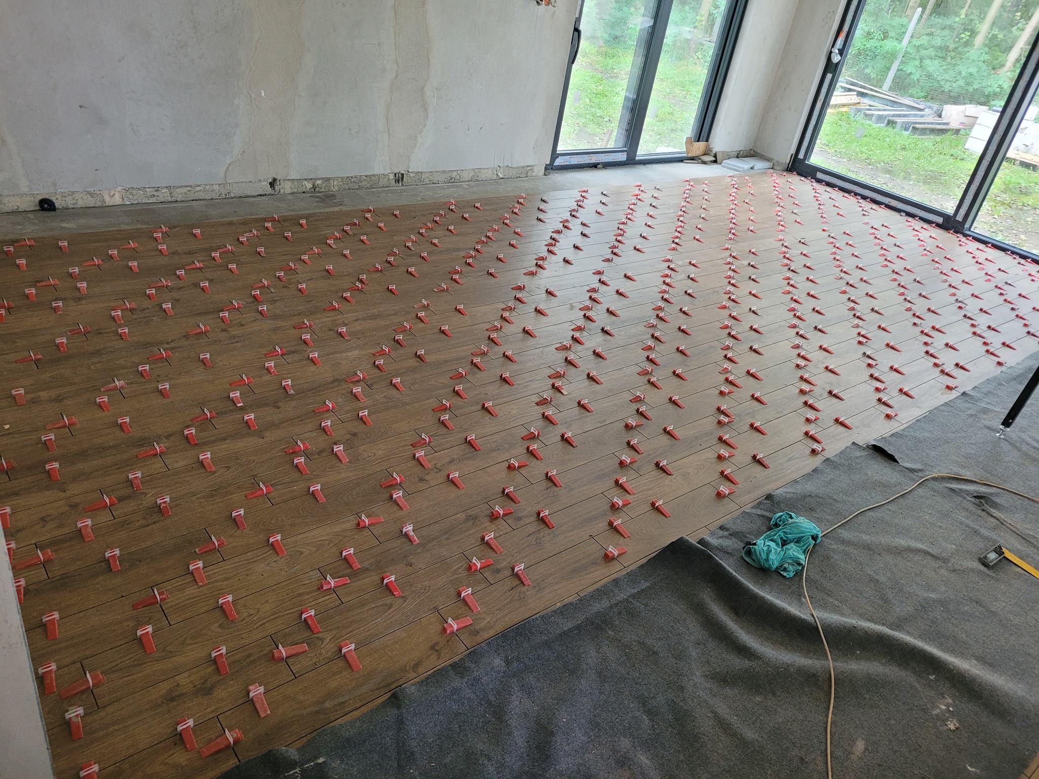 Układanie brązowych płytek podłogowych imitujących drewno, z zastosowaniem czerwonych klinów poziomujących, widoczne okno z widokiem na zieleń, szara mata ochronna i poziomica.