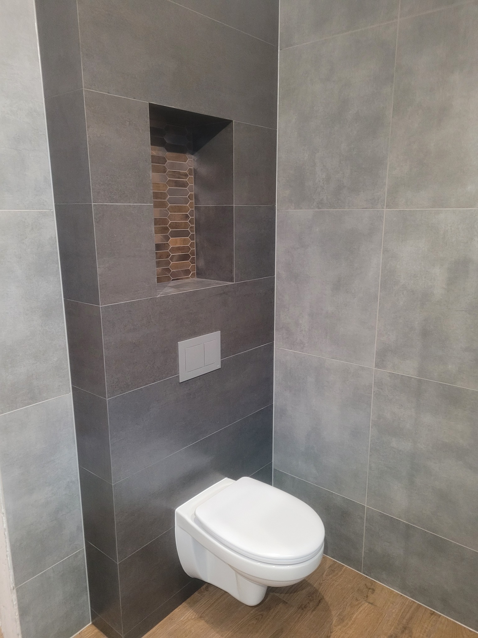 Nowoczesna toaleta podwieszana z szarą, wielkoformatową glazurą, drewnopodobna podłoga, wnęka z dekoracyjną mozaiką w ścianie.