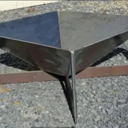 Surowa, metalowa konstrukcja w kształcie odwróconej piramidy, spawana z blachy i profili, stojąca na żwirowym podłożu.