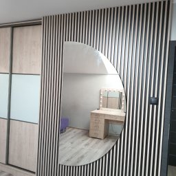 ARMAR MEBLE - Ściana z pionowych lameli w kolorze czarno-beżowym z owalnym lustrem, w którym odbija się toaletka z oświetleniem i fioletowy puf, obok szafa przesuwna z drewnianymi i szklanymi panelami,...
