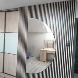 Ściana z pionowych lameli w kolorze czarno-beżowym z owalnym lustrem, w którym odbija się toaletka z oświetleniem i fioletowy puf, obok szafa przesuwna z drewnianymi i szklanymi panelami,...