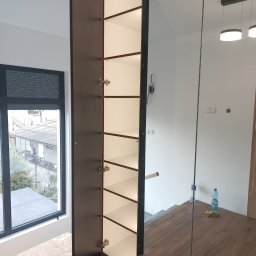 ARMAR MEBLE - Wysoka szafa wnękowa z ciemnobrązowymi drzwiami i białym wnętrzem, jedna strona otwarta, widok na okno z roletami zewnętrznymi i narzędzia na podłodze, odbicie w lustrzanej części szafy.