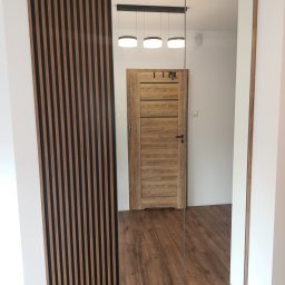 ARMAR MEBLE - Nowoczesne wnętrze z drewnianą ścianką działową z lameli i lustrzaną szafą w przedpokoju; widoczna drewniana podłoga i drzwi wewnętrzne.
