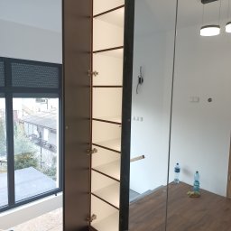 ARMAR MEBLE - Wysoka, otwarta szafa z ciemnego drewna z jasnymi półkami, widok z góry, fragment okna z roletami w tle, wnętrze w trakcie wykończenia.