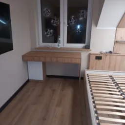 Drewniane biurko z szufladami i szafką obok okna z dekoracjami świątecznymi, zintegrowane z zabudową meblową pod skosem dachu, obok rama łóżka bez materaca.