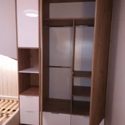 Otwarta szafa wnękowa z drewnianymi elementami i białymi frontami szuflad, widoczny stelaż łóżka w tle.