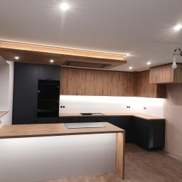 ARMAR MEBLE - Nowoczesna kuchnia z wyspą, fronty w kolorze grafitowym i drewnopodobnym, oświetlenie LED podszafkowe i sufitowe, widoczne narzędzia i materiały budowlane w oknie.