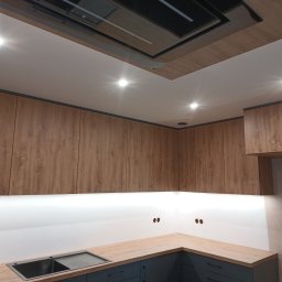 ARMAR MEBLE - Nowoczesna kuchnia z drewnianymi szafkami górnymi, szarymi szafkami dolnymi i oświetleniem LED pod szafkami. Widoczne gniazdka elektryczne na ścianie.