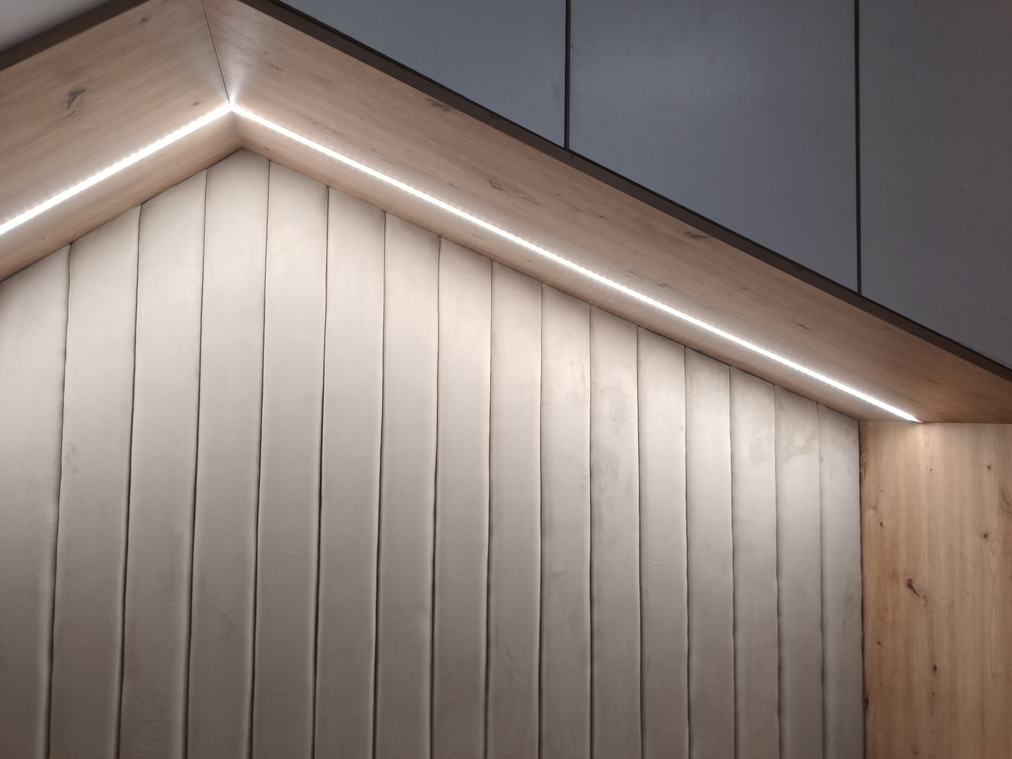 Panel tapicerowany w pionowe pasy, obudowany drewnem i oświetlony listwą LED, tworzący wnękę.