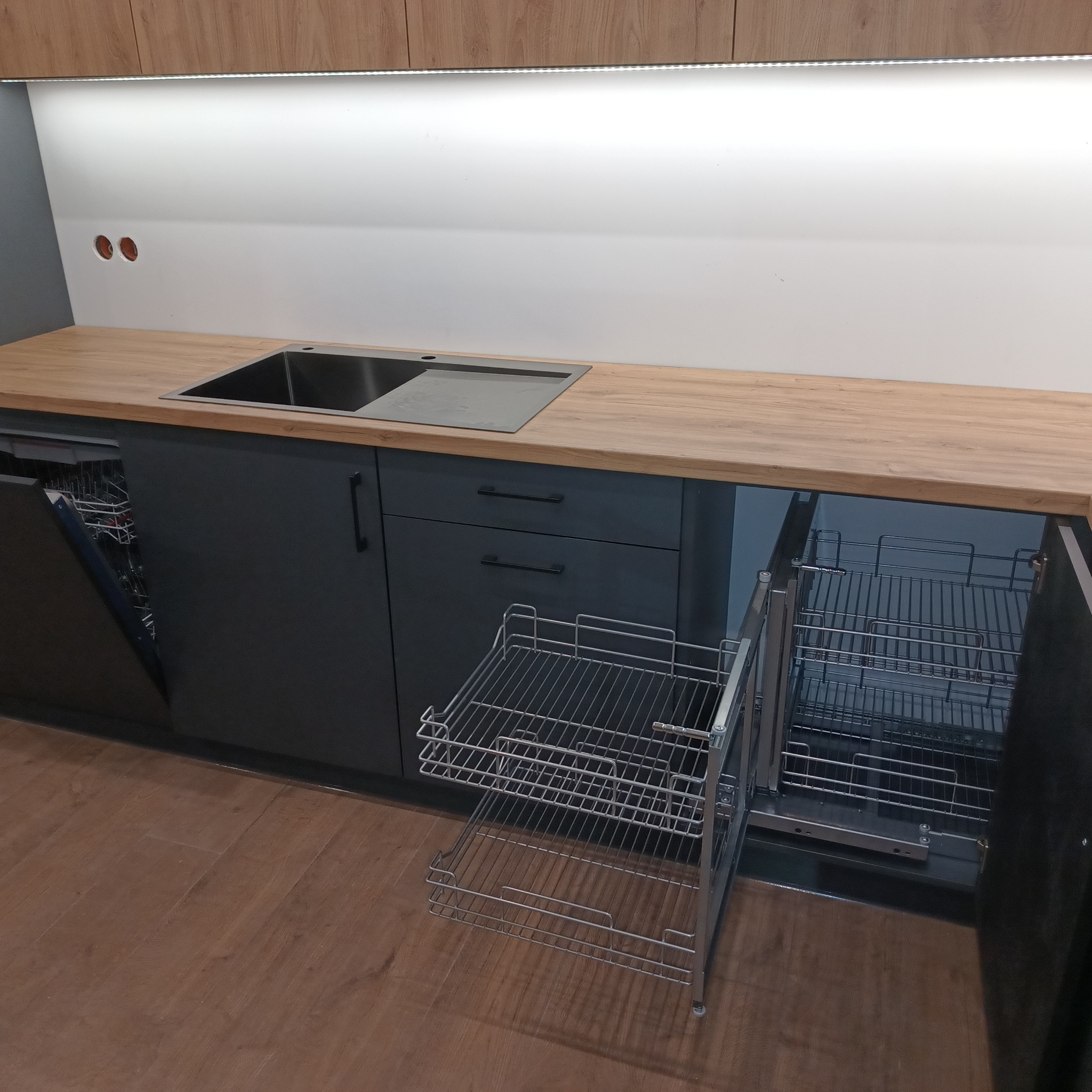 Szare szafki kuchenne z drewnianym blatem, wysuwany kosz cargo na naczynia i otwartą zmywarką po lewej stronie.