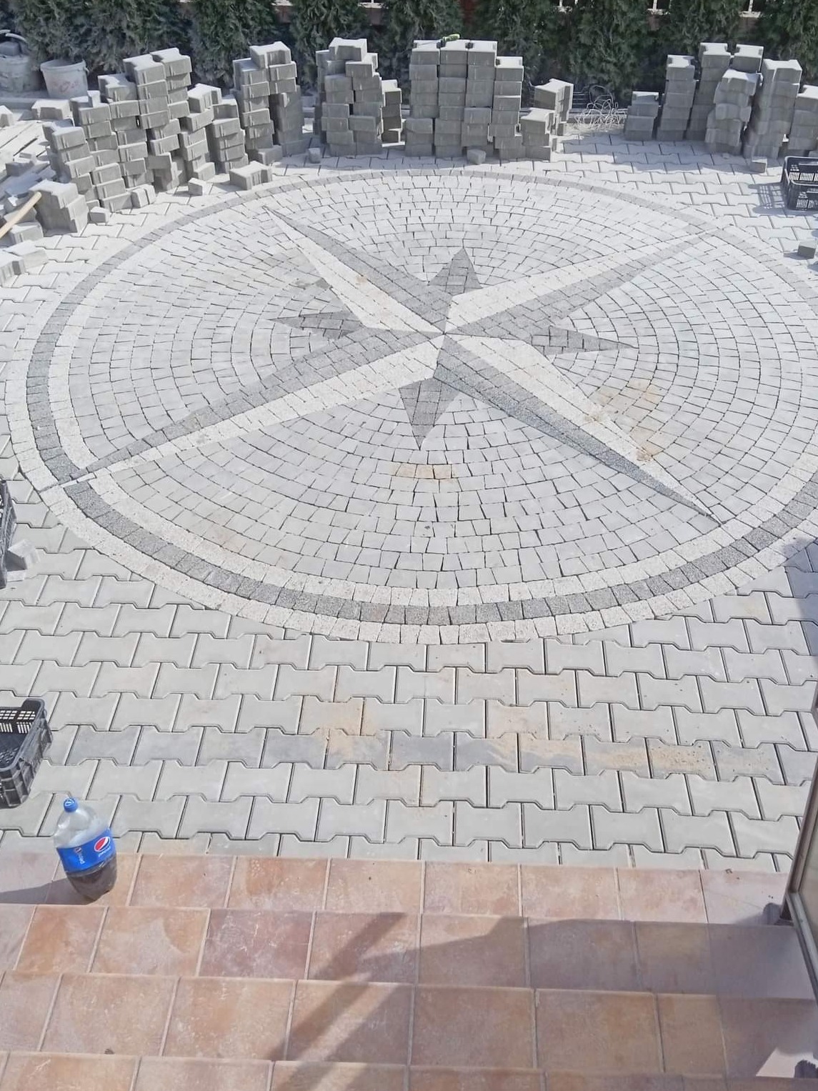 Okrągły plac z szarej kostki brukowej ułożonej w wzór kompasu, otoczony stosem nieużytej kostki, widok z góry.