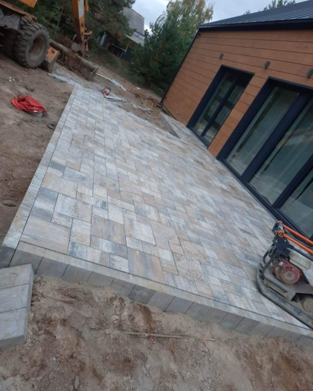 Ułożona kostka brukowa w odcieniach szarości i beżu przy nowoczesnym domu z drewnianą elewacją, widoczna ubijarka wibracyjna i ciężki sprzęt budowlany w tle.