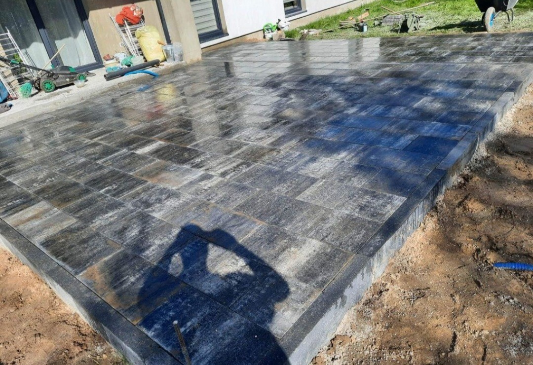 Ułożona, mokra kostka brukowa w odcieniach szarości i czerni, widoczne narzędzia i materiały budowlane w tle, cień fotografa w dolnej części zdjęcia.