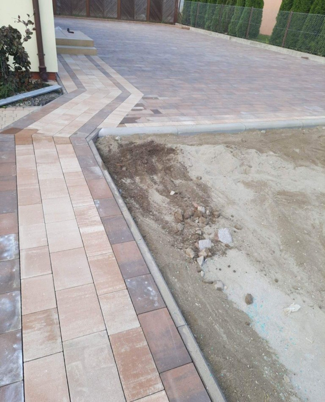 Ułożona kostka brukowa w odcieniach brązu i beżu, z dekoracyjnym wzorem, obok przygotowany teren z piaskiem i gruzem pod dalsze prace.