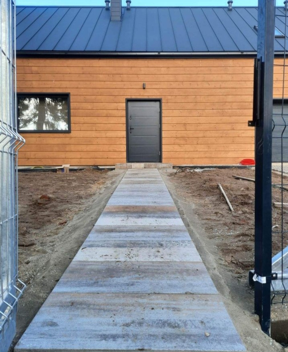 Ścieżka z szarych płyt prowadząca do drewnianego domu z czarnymi drzwiami i dachem, widok zza ogrodzenia panelowego.