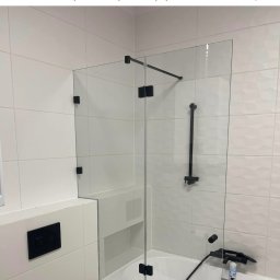 HomeGlass - Szklana kabina prysznicowa z czarnymi okuciami zamontowana nad wanną w łazience wyłożonej białymi, wzorzystymi płytkami.