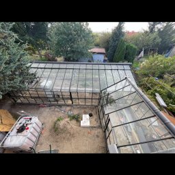 HomeGlass - Widok z góry na szklaną konstrukcję oranżerii z czarną ramą, wewnątrz widoczne przygotowane podłoże, obok pojemnik z cegłami i narzędzia, otoczenie to ogród z drzewami.