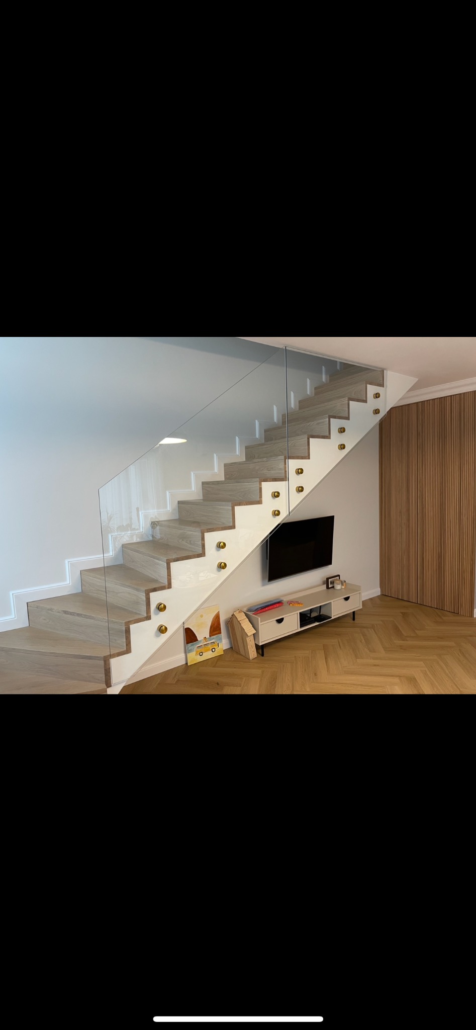 Schody drewniane z balustradą szklaną mocowaną na złotych uchwytach, widok na salon z parkietem w jodełkę i telewizorem.
