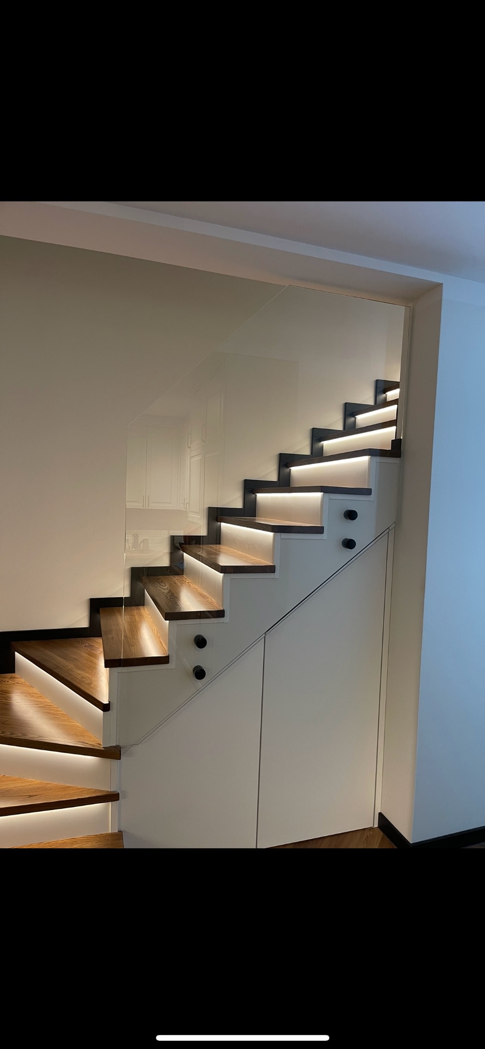 Nowoczesne schody z drewnianymi stopniami podświetlonymi LED, zabezpieczone szklaną balustradą, zintegrowane z białą zabudową meblową pod schodami.