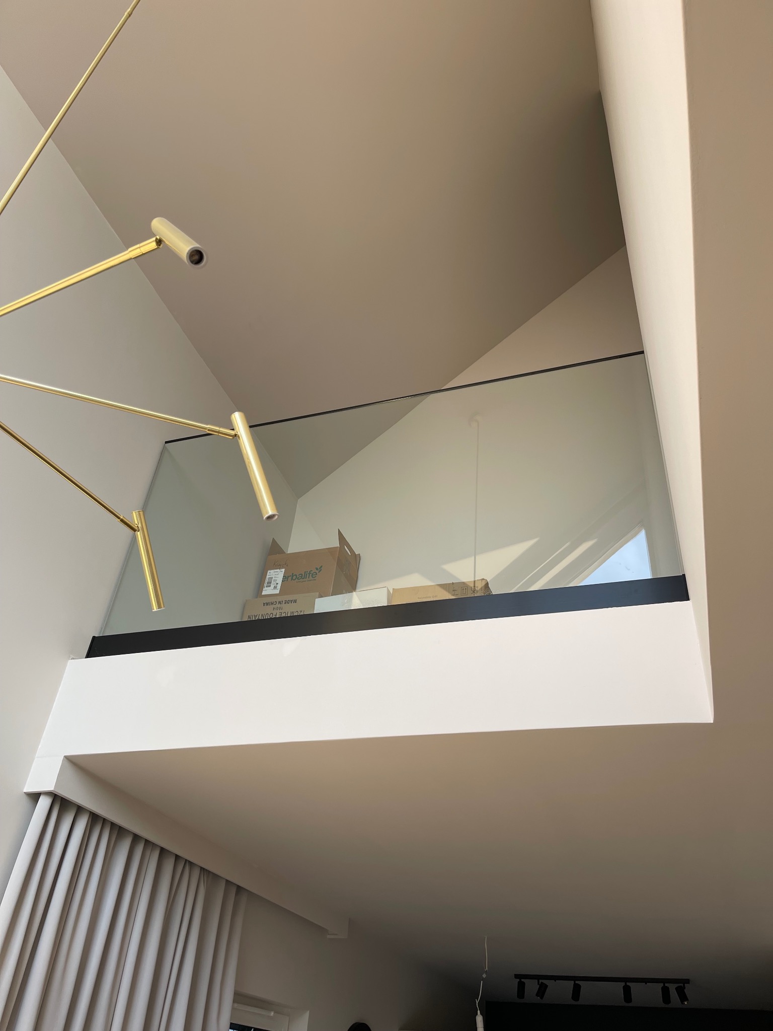 Szklana balustrada na antresoli z czarnym profilem, widok z dołu, z widoczną częścią złotego żyrandola i kartonami na antresoli.