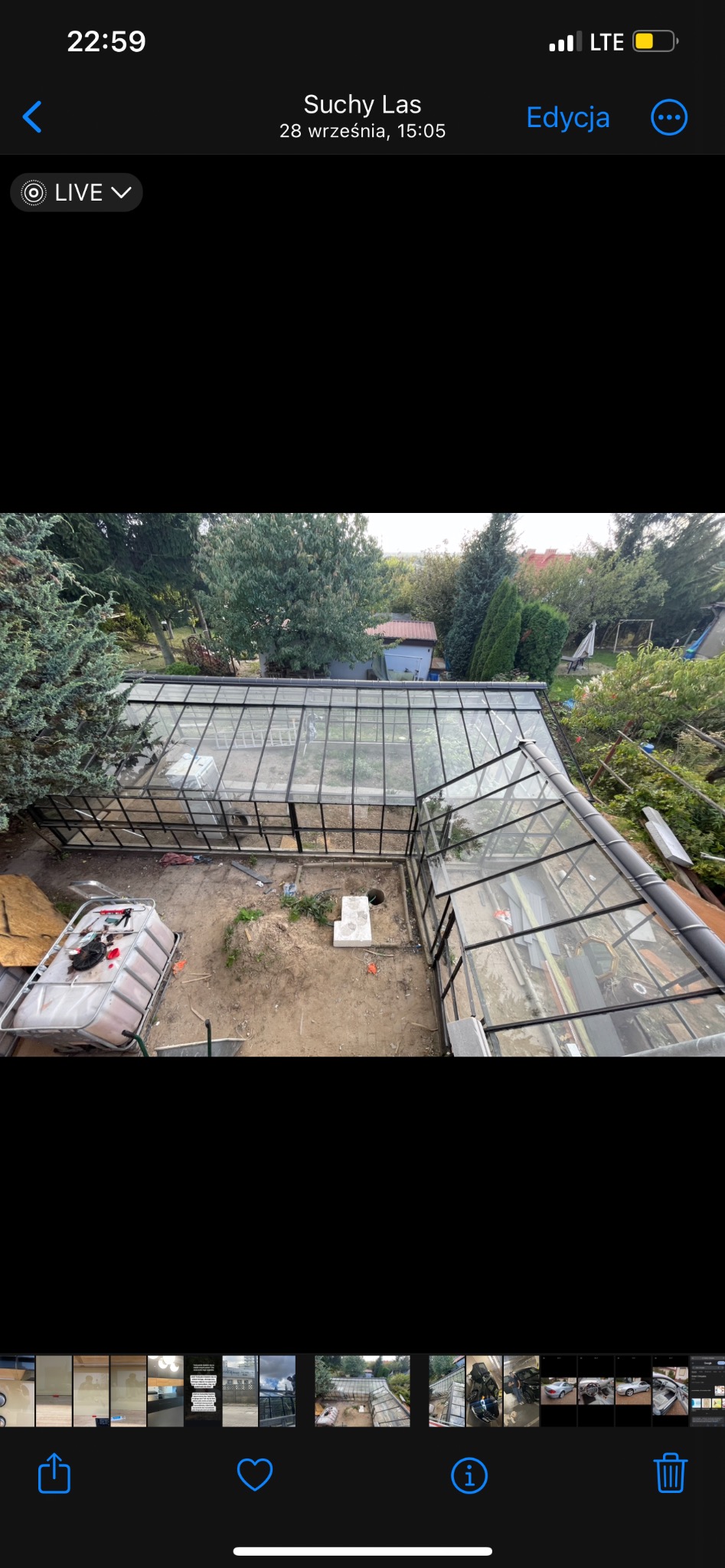 Widok z góry na szklaną konstrukcję oranżerii z czarną ramą, wewnątrz widoczne przygotowane podłoże, obok pojemnik z cegłami i narzędzia, otoczenie to ogród z drzewami.