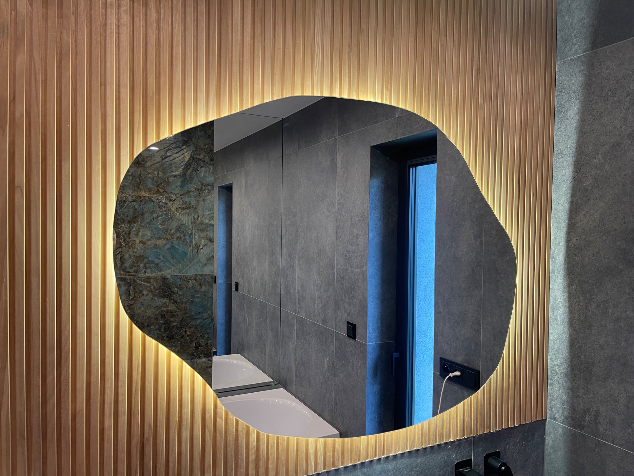 Nieregularny kształt podświetlanego lustra łazienkowego w drewnianej ramie z pionowych listewek, odbijającego fragment ściany z szarych płytek i drzwi.