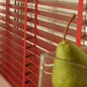 Czerwone drewniane żaluzje poziome z widoczną taśmą podtrzymującą, częściowo zasłonięte przez szklankę z zieloną gruszką.