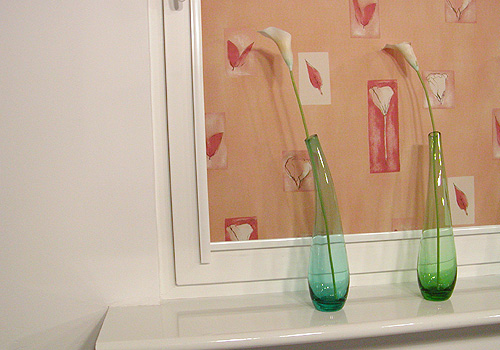 Dwa smukłe, szklane wazony z kaliami stojące na białym parapecie okiennym, z tapetą w tle w odcieniach różu i brzoskwini z motywem liści i kwiatów.