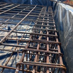 Kam-Bud - Siatka zbrojeniowa ułożona na czarnej folii hydroizolacyjnej, pionowe pręty zbrojeniowe wystają ponad siatkę, widoczne elementy mocujące pręty, w tle fragment dachu budynku.