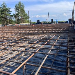 Kam-Bud - Siatka zbrojeniowa ułożona na czarnej folii, widok z góry na plac budowy z drzewami w tle i niebem z chmurami.