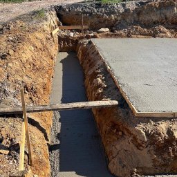 Kam-Bud - Wylana betonowa podstawa fundamentu w wykopie, z widocznym deskowaniem i drewnianymi podporami, teren budowy.