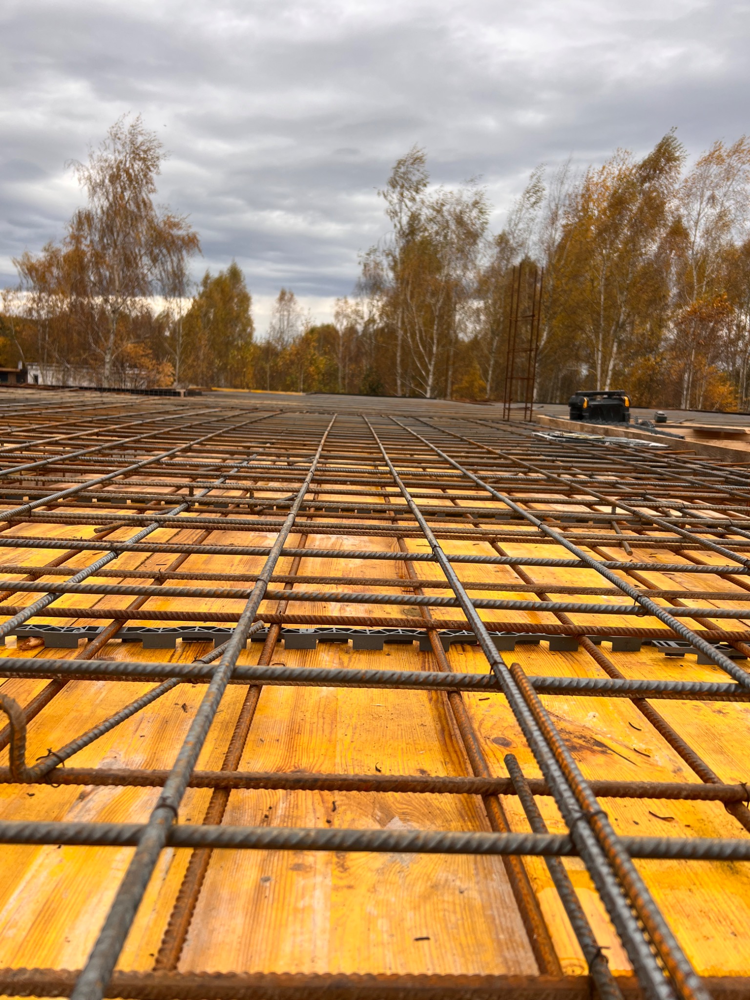 Siatka zbrojeniowa ułożona na żółtym szalunku, widok z dołu pod kątem, w tle drzewa i pochmurne niebo.