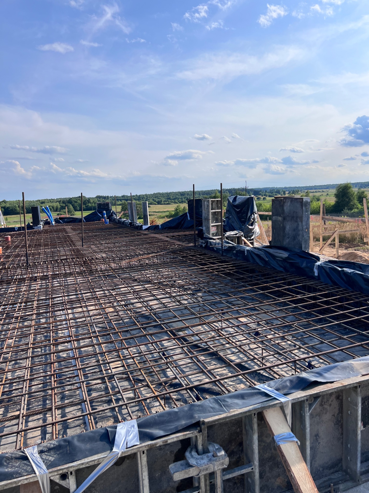 Siatka zbrojeniowa ułożona na deskowaniu fundamentów, widoczne elementy szalunków, folia ochronna i wystające pręty zbrojeniowe, w tle krajobraz z drzewami i błękitne niebo.