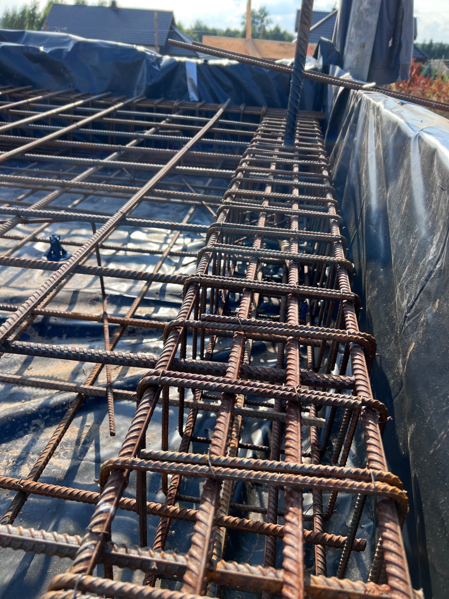 Siatka zbrojeniowa ułożona na czarnej folii hydroizolacyjnej, pionowe pręty zbrojeniowe wystają ponad siatkę, widoczne elementy mocujące pręty, w tle fragment dachu budynku.
