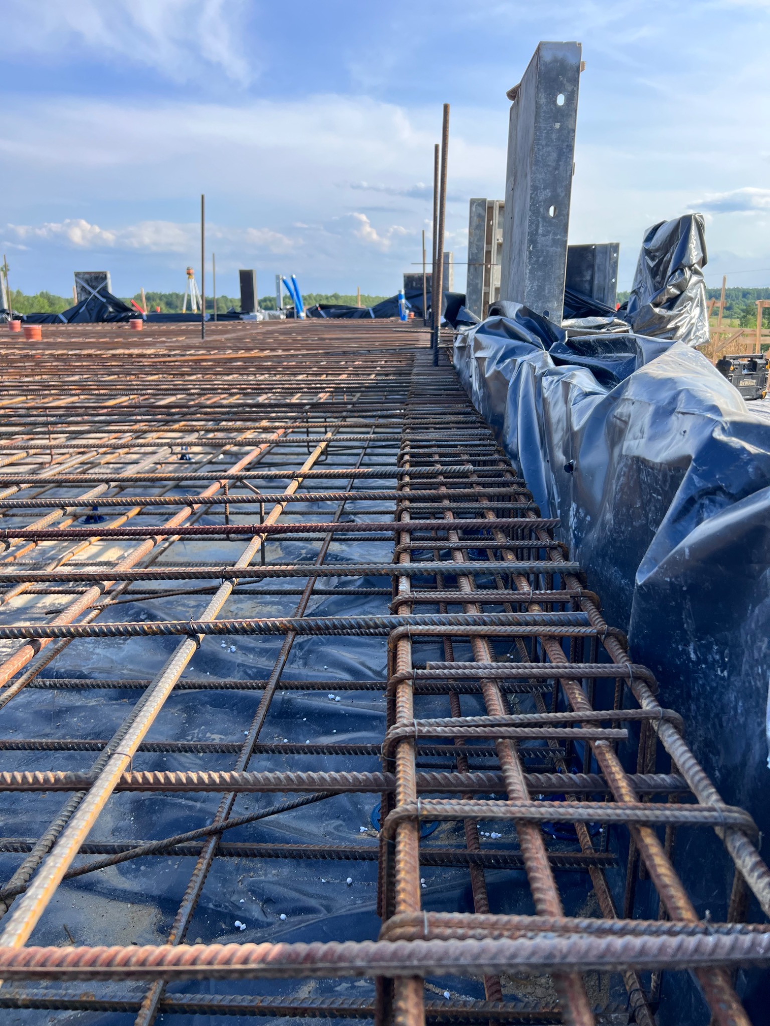 Siatka zbrojeniowa ułożona na czarnej folii, przygotowana do wylewki fundamentów, widok z niskiej perspektywy, w tle niebo z chmurami.