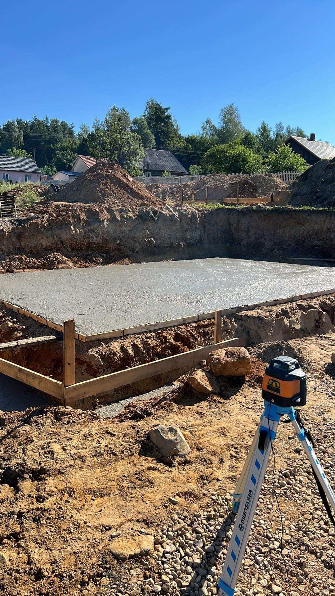 Świeżo wylana, gładka płyta fundamentowa w drewnianym szalunku, z widocznym niwelatorem laserowym na statywie, otoczona wykopem i ziemią.