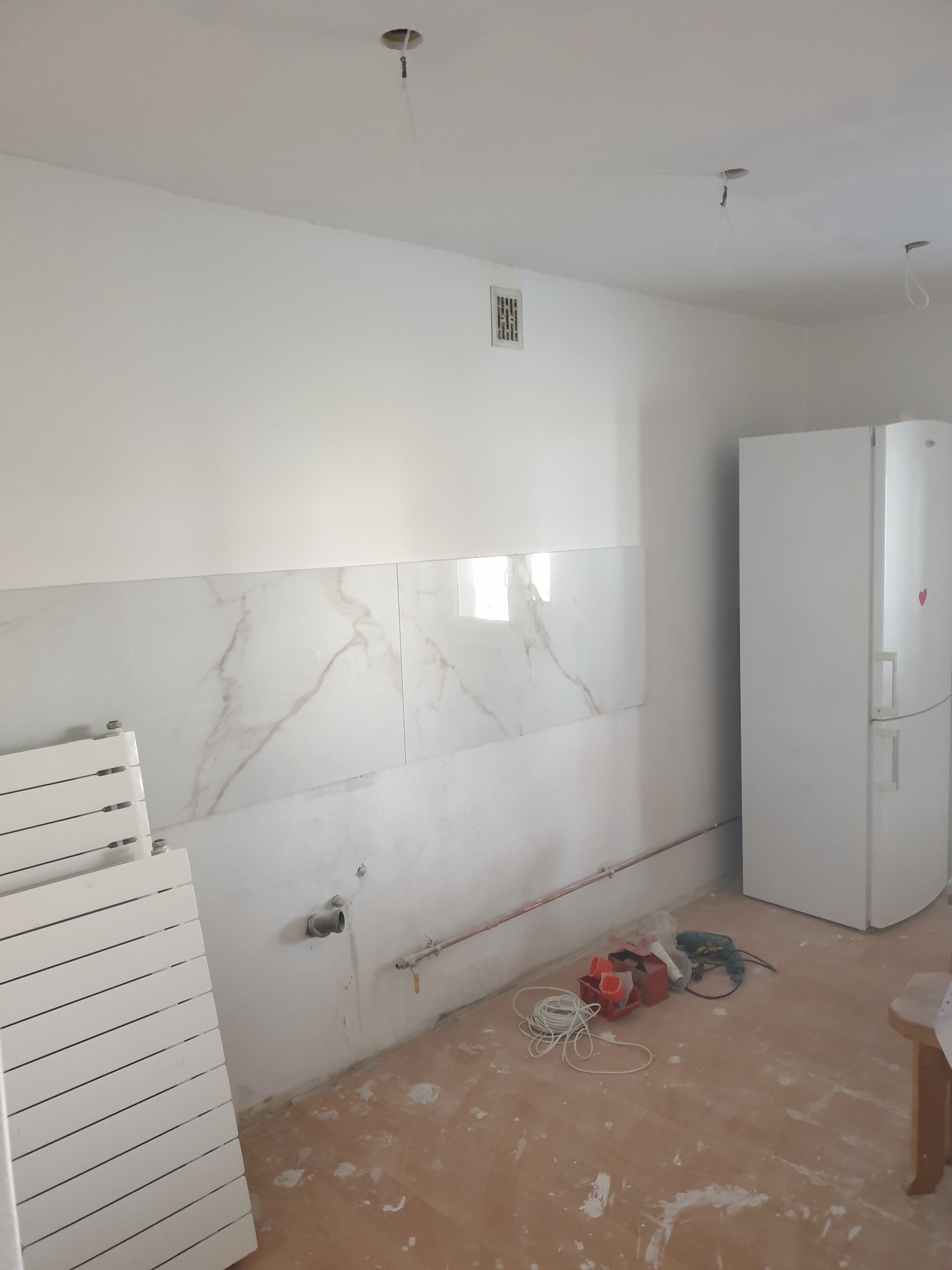 Kuchnia w trakcie remontu: położone marmurowe płytki nad blatem, biała lodówka, przygotowane do montażu białe panele grzejnikowe, narzędzia i przewody na podłodze, widoczne instalacje.