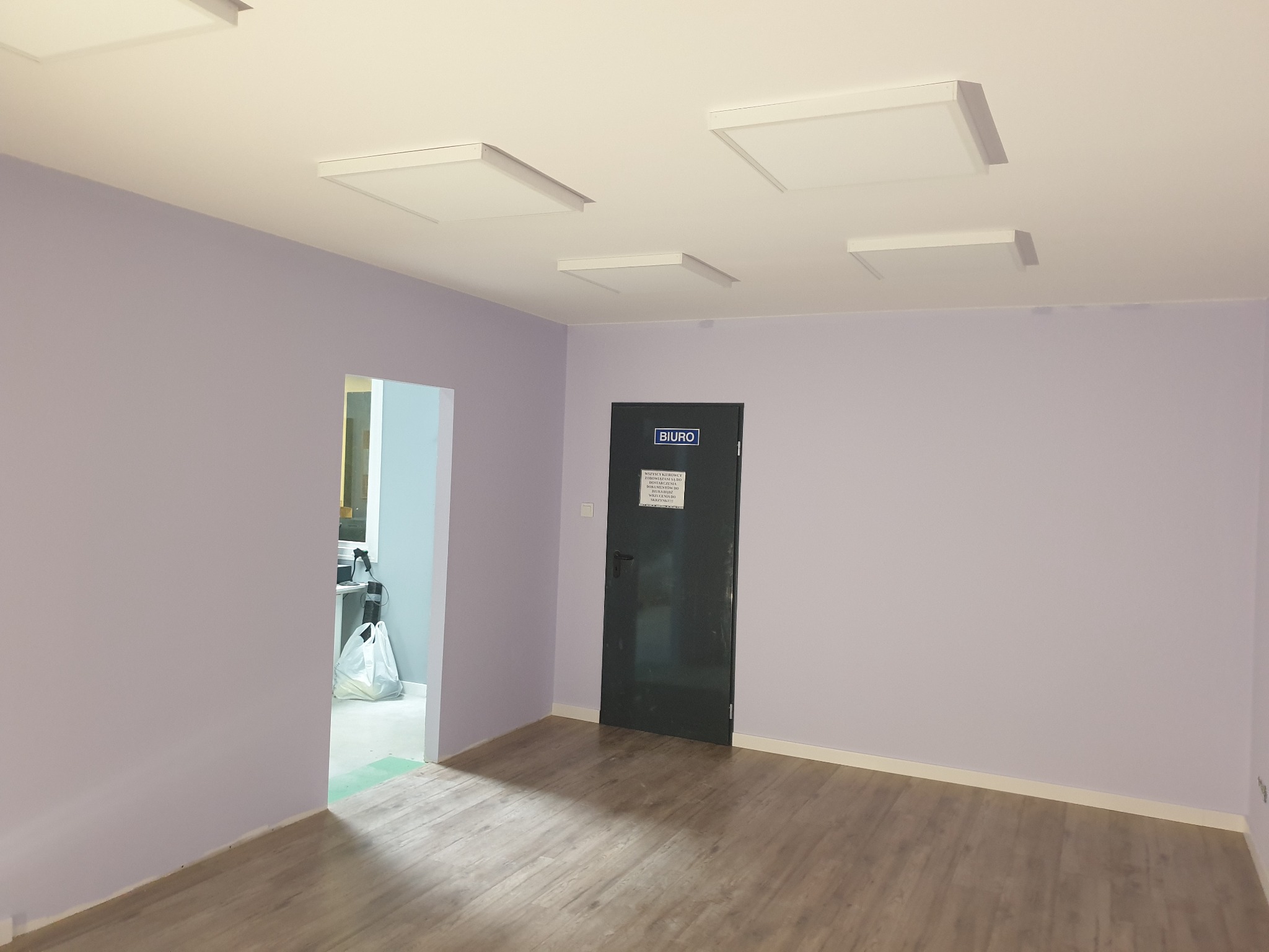 Wnętrze biura po malowaniu, ściany w odcieniach fioletu, drewnopodobna podłoga, białe panele sufitowe i czarne drzwi z napisem 'BIURO'.