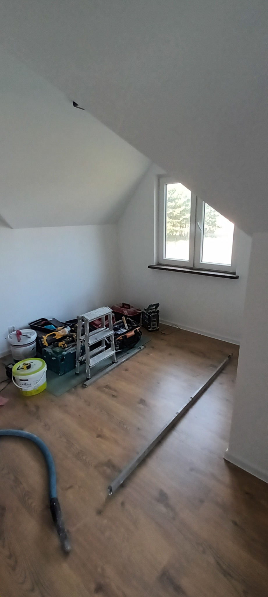 Poddasze po remoncie z nową podłogą z paneli; widoczne narzędzia, drabina, poziomica i wiadro farby.