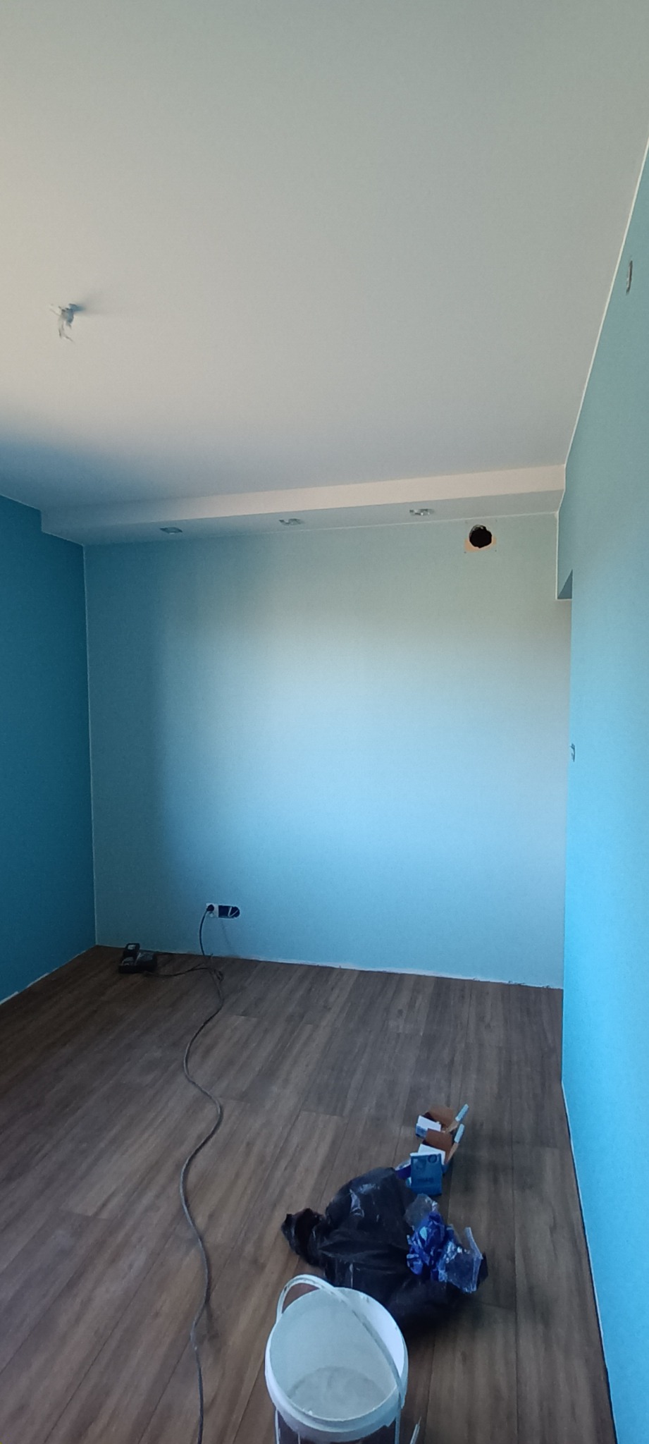 Pokój po malowaniu ścian na dwa odcienie błękitu, z widoczną podłogą z paneli, puszką po farbie, workiem na śmieci i gniazdkiem elektrycznym z podłączonym kablem.